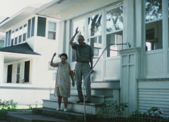 An elderly couple wave hello from their front stoop