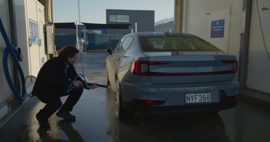 A man is standing outside at a car wash washing his car with a hose