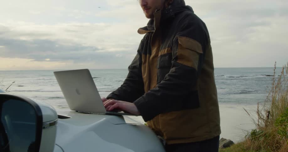 A man is standing at the beach typing on his computer that is sitting on his vehicle