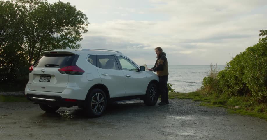 A man is parked along the coast and standing outside typing on his laptop computer on the hood of his car