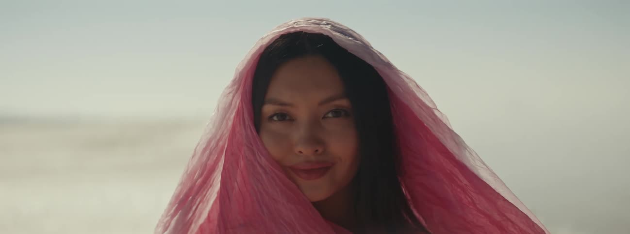 A woman with a sheer pink veil over her head smiles and blinks