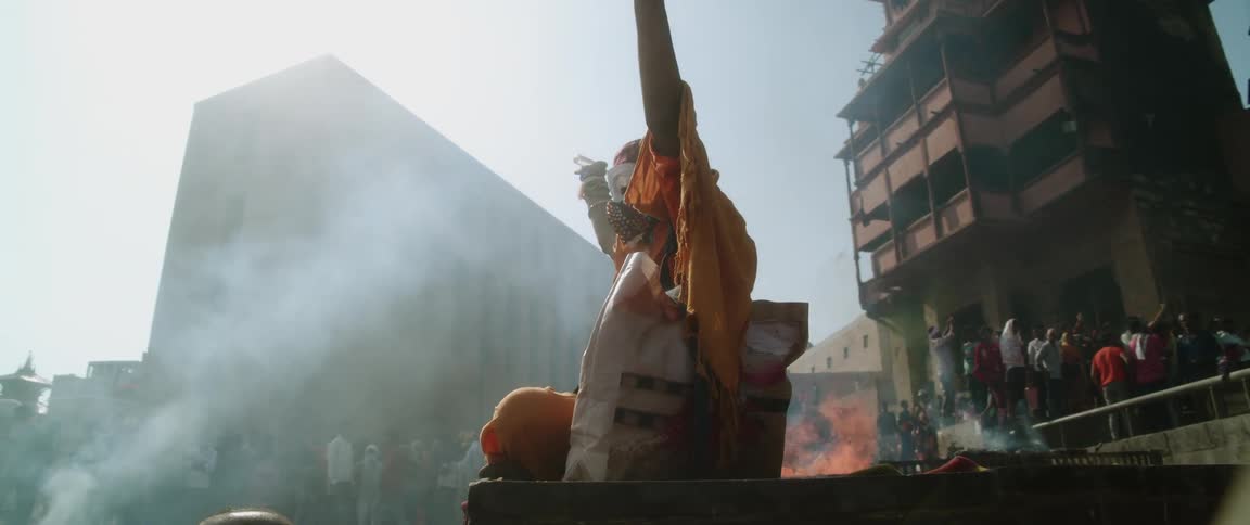 A woman sits on a stage with a large crowd of people and raises her arms in the air as fire burns behind her