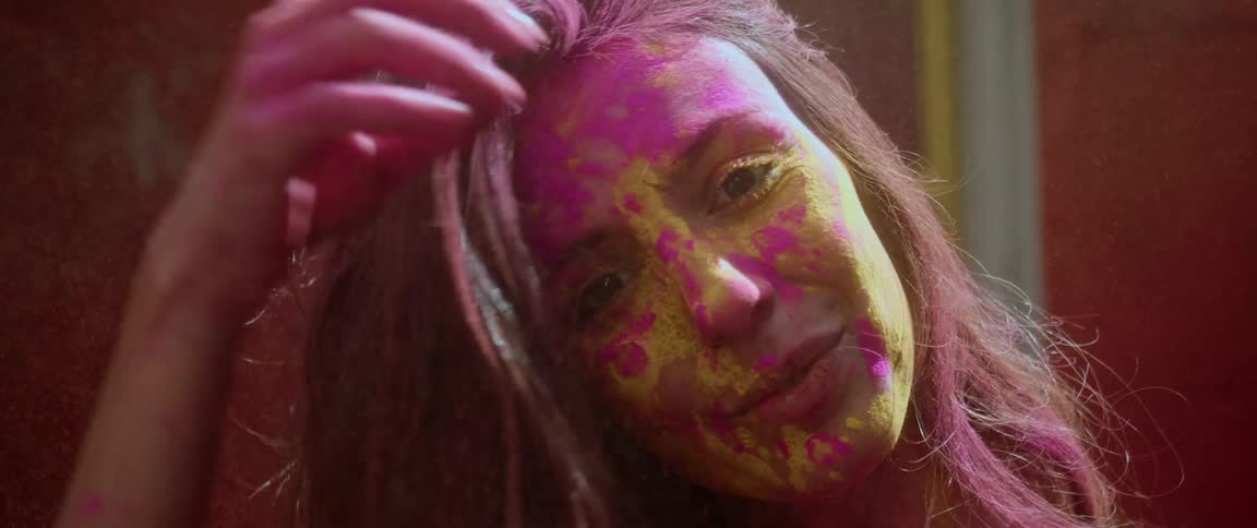 A teenage girl is standing outside smiling with pink paint on her face as she pulls back her hair with her hand