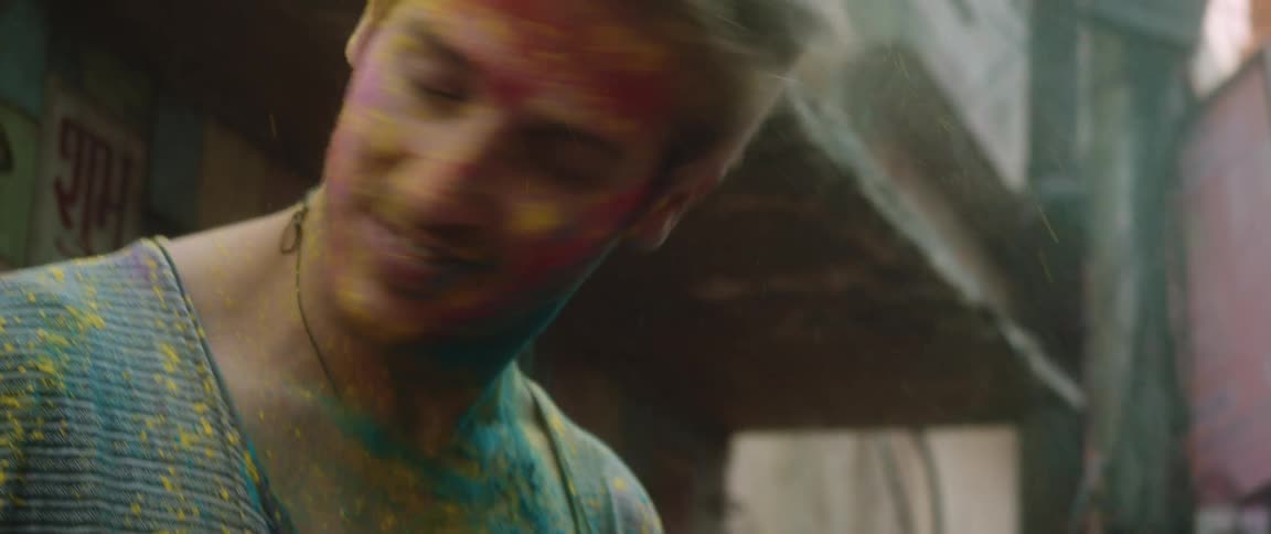 A man with red paint on his face is standing on the street shaking his head as blue and yellow powder falls from his hair