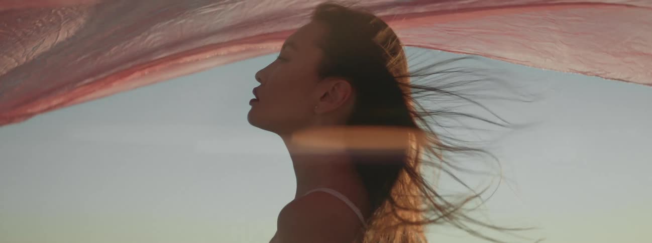 A woman with her eyes closed lets the wind shake a sheer pink veil over her head
