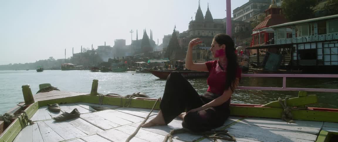 Womanwith red powder coated face sits on boat moving through water just off coast near narrow domed buildings