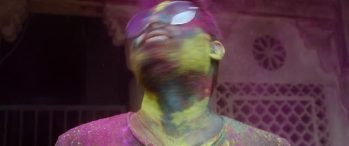 A man with his beard sunglasses face and hair covered in powdered dye shakes his head and spreads the powder all around him as he smiles