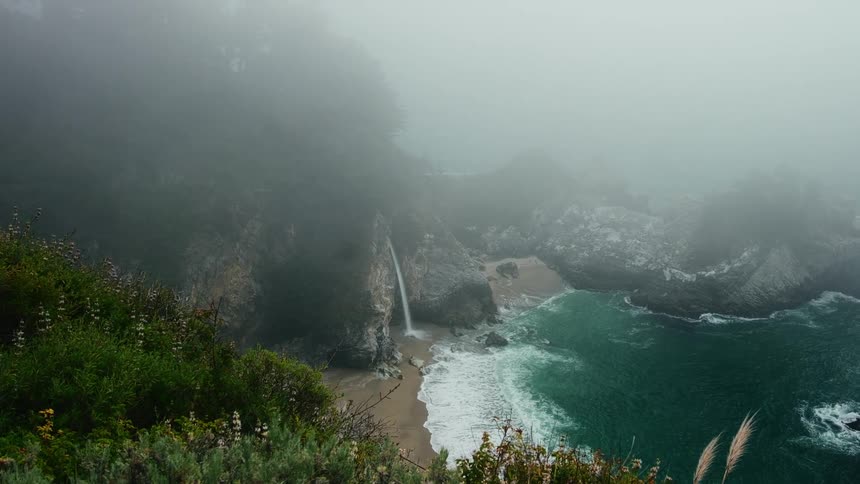 Looking over a mountain as waves roll into shore and over a small beach and water falls in front of a sea cave under a foggy sky
