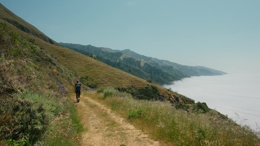 A woman hiking on a dirt path between grassy hills and the ocean