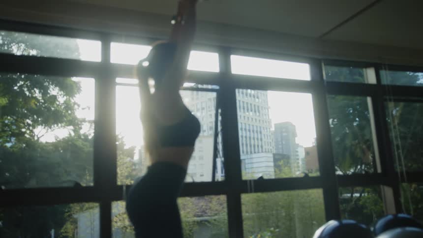 Woman lifts weight over her head in gym near window showing city buildings