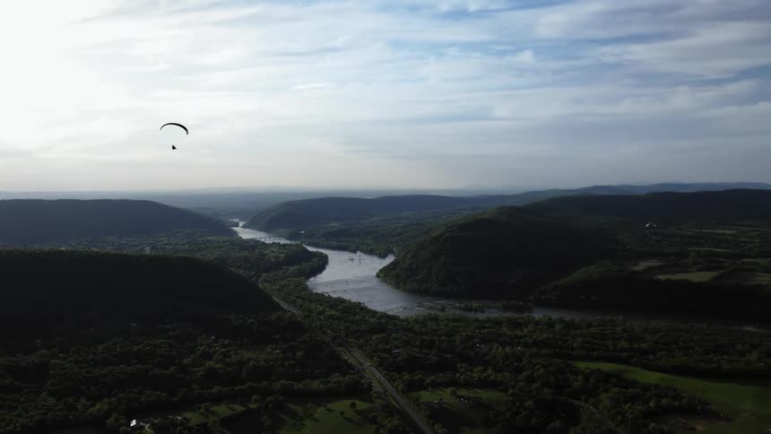 A paraglider flies over a mountain river valley under a cloudy sky