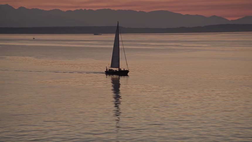Aerial footage of a sailboat gracefully sailing through Elliott Bay at sunset showcasing the beautiful scenery of Seattle Washington with a vibrant red sky in the background