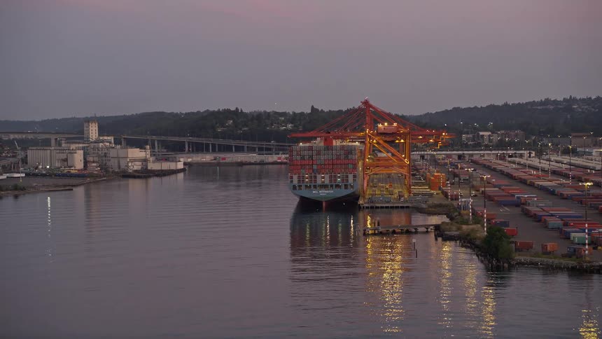  aerial footage of a cargo ship docked at the Port of Seattle during sunset with cranes loading containers onto it