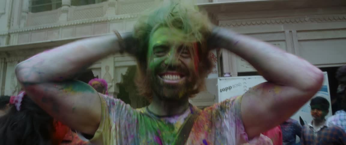 A man with his face hair beard and clothes covered in powered dye rubs and shakes his hair spreading the powder then raises his arms in celebration while smiling