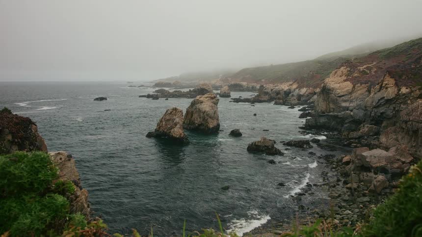 An aerial of ocean waves crashing against the rocky coastline on a foggy morning