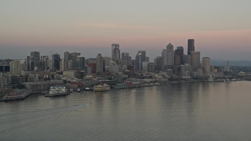  aerial footage of the Central Waterfront and Downtown Seattle skyline at sunset showcasing the city's high-rise buildings and bustling shoreline as a motorboat glides past the docks