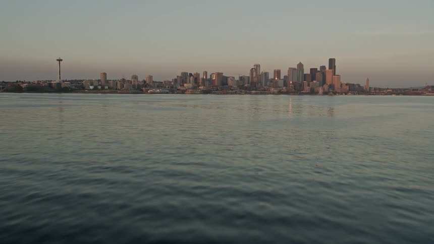 an aerial view of the Seattle Space Needle and downtown skyline at sunset as the camera tilts up from Elliott Bay revealing the iconic structure amidst towering buildings and the picturesque waterfront
