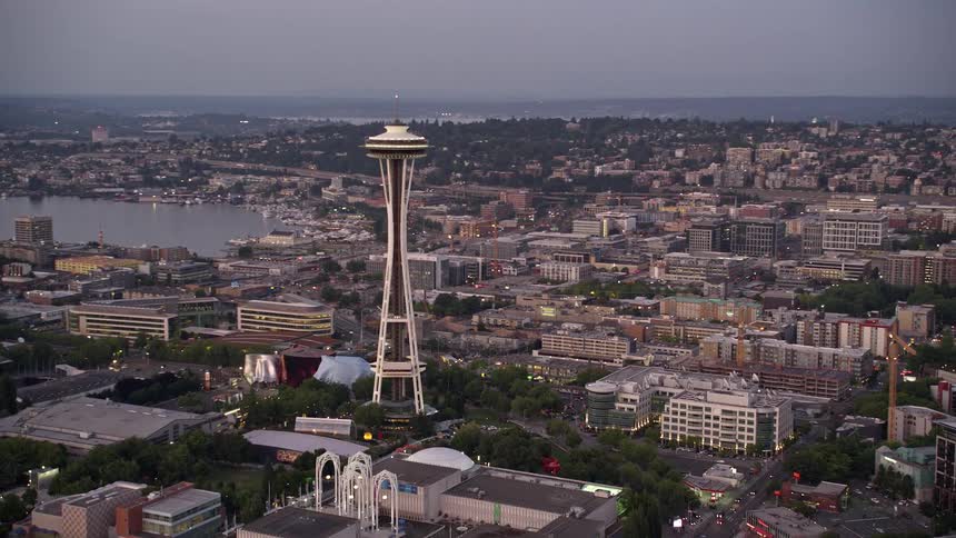 Aerial footage captures the iconic Space Needle towering over buildings as it tilts up from Elliott Bay to reveal the Seattle Central Waterfront piers in Downtown Seattle during a misty sunset