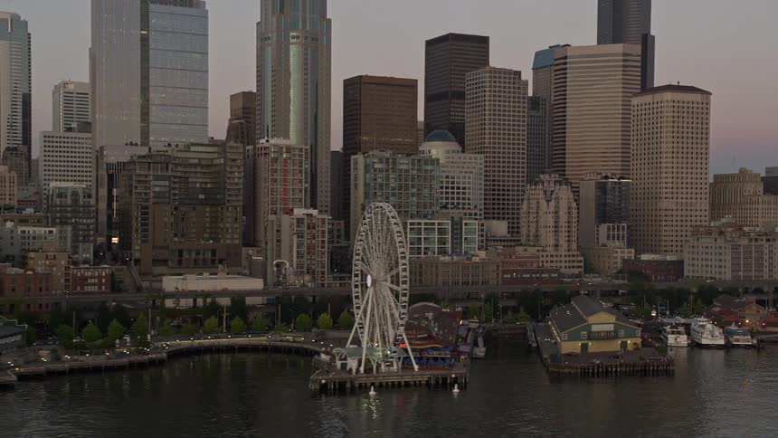 Low approach to the Great Wheel and Waterfront with the towering skyline of Downtown Seattle during sunset as seen from an aerial perspective