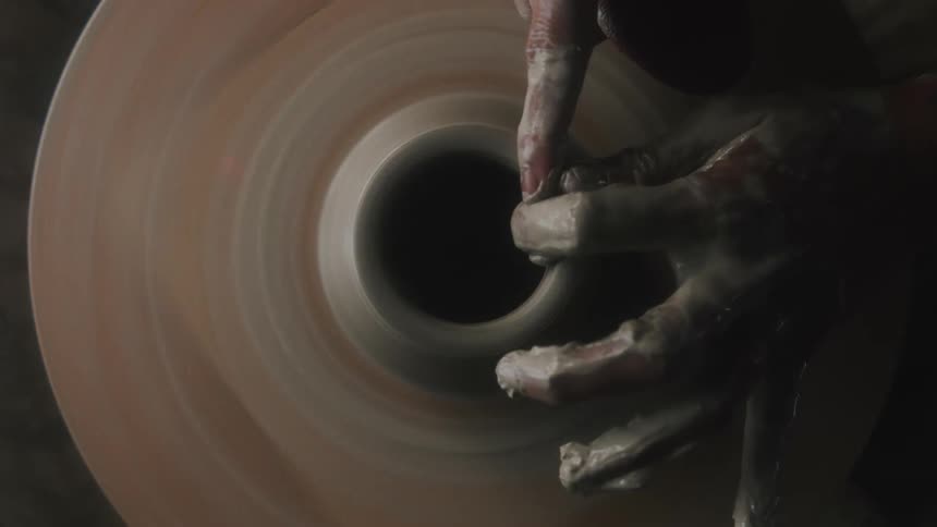 Person uses clay smeared hands to form shape spinning on pottery wheel
