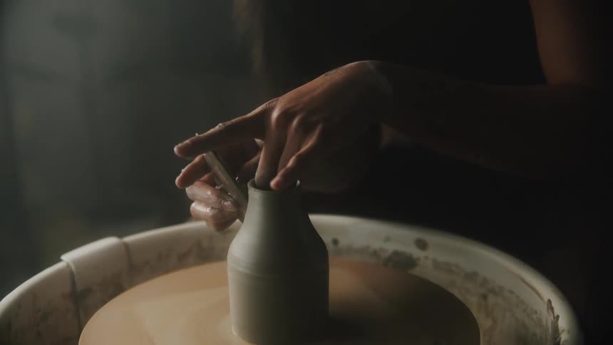 Woman sits and shapes vessel on spinning pottery wheel