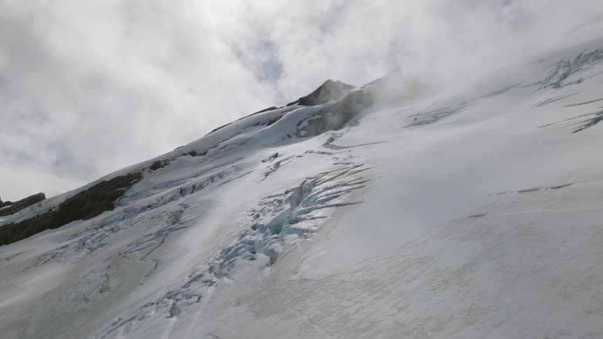 Snow covering glacial mountainside contains cracks in places under an overcast sky