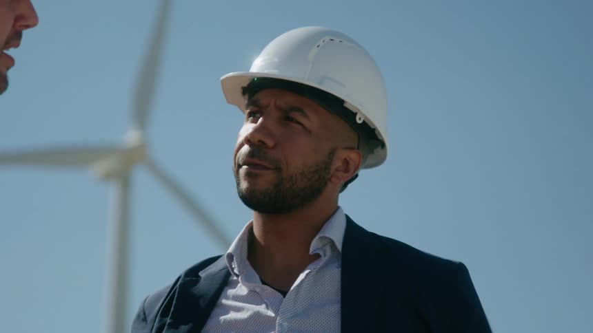 Engineers with hard hats and jackets talk with each other with wind turbine in the background