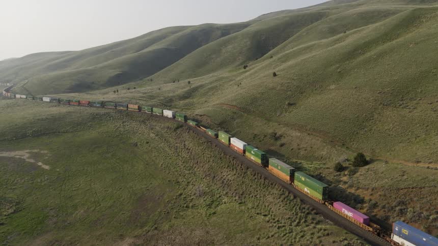 A very long container train travels along lush painted hills in morning light