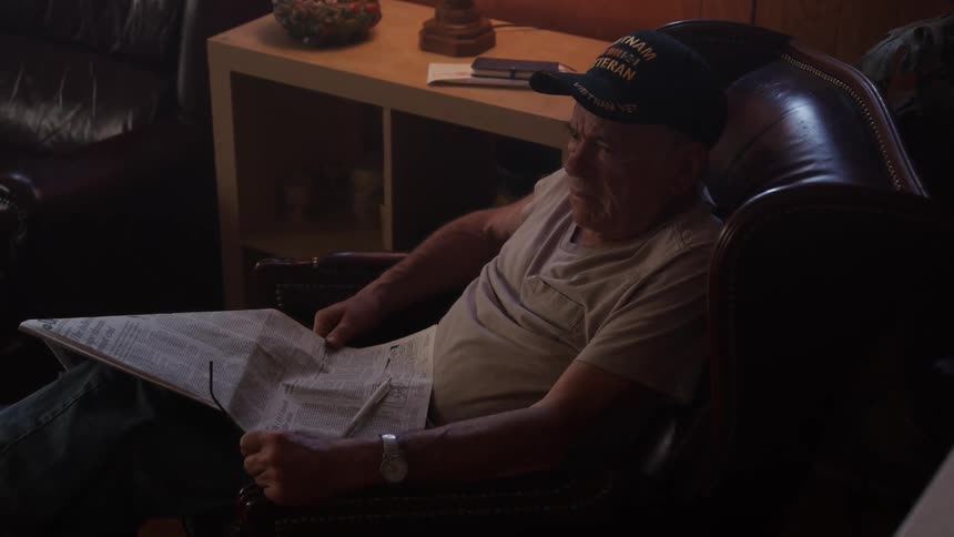 A man reads a newspaper in his chair as another person walks by him