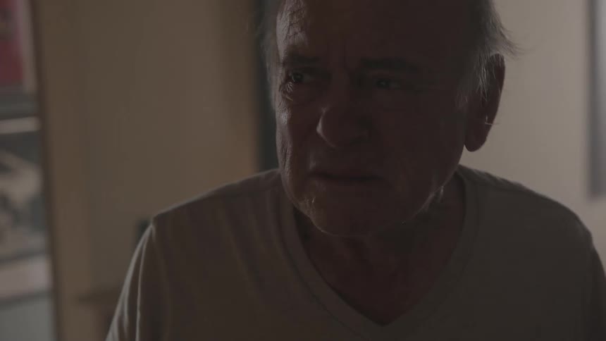 An elderly man is standing in his house looking angry and sad