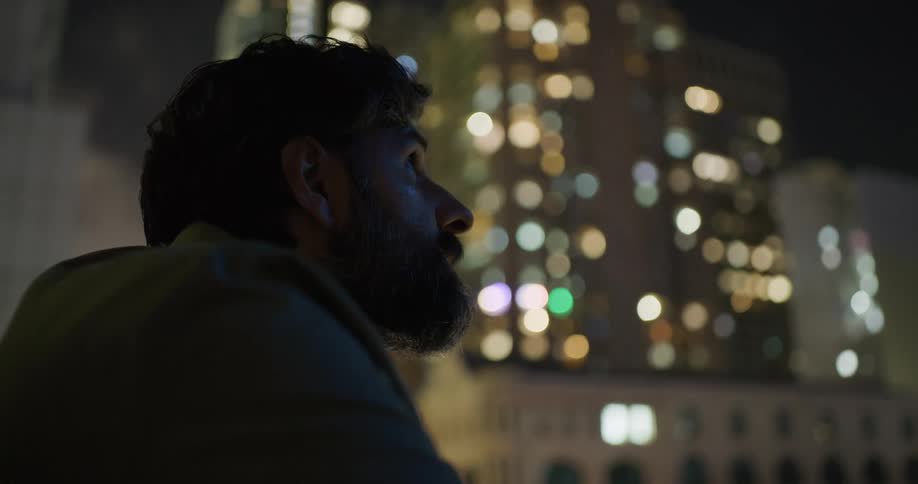 With illuminated skyscrapers in the background man with beard and jacket looks around