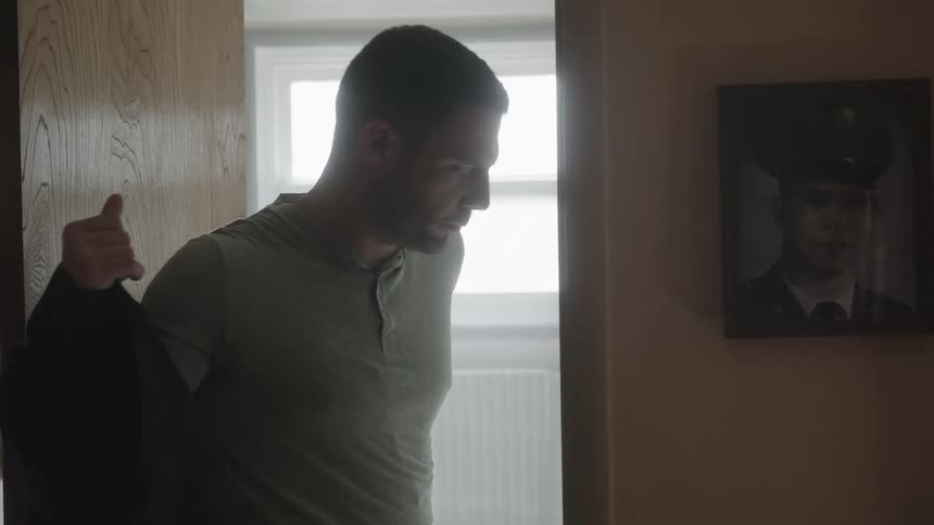 Man smooths his hair in bathroom then dons jacket near wall with picture of man in uniform