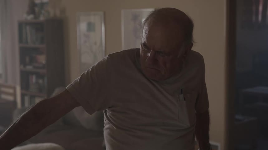 An old man sporting an angry frown paces around a living room looking pensive