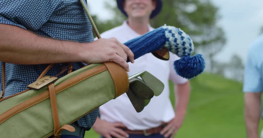 A man holds a bag of golf clubs with wool protectors over them while another man smiles at him with hands on his hips
