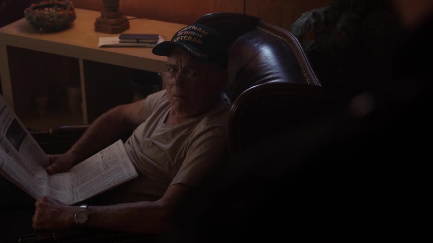 A man reading a newspaper speaks with another person in the living room