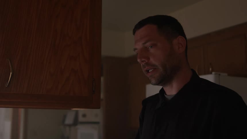 A man is standing in a kitchen talking to someone else and holding a pen
