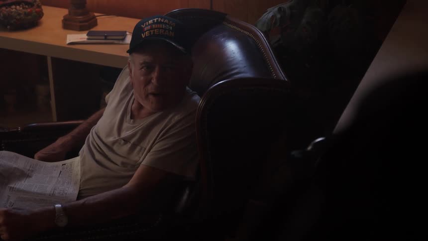 An elderly man sitting in a lounge chair wearing a hat and with a newspaper on his lap speaks angrily to someone else in the room