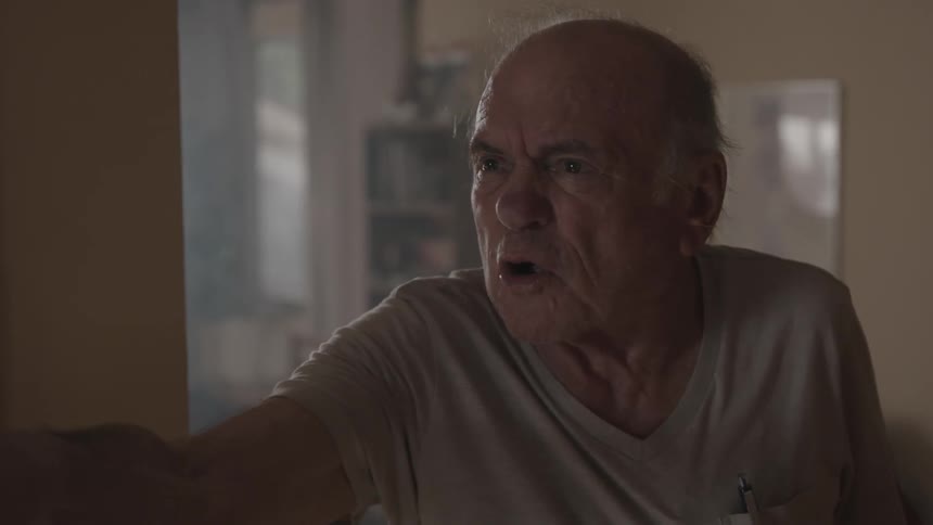An elderly man turns and shouts angrily at someone holding up a pen