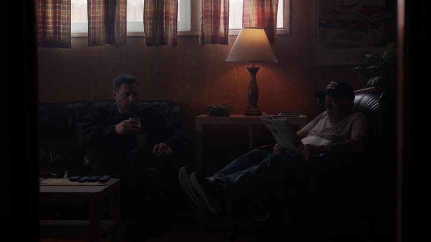 A man walks into the living room with a drink in his hand and sits down on the couch next to another man in a chair
