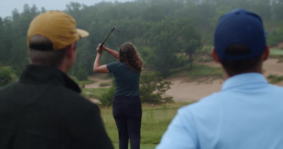 A woman takes a strike at a golf ball swinging with her club as two man watch her