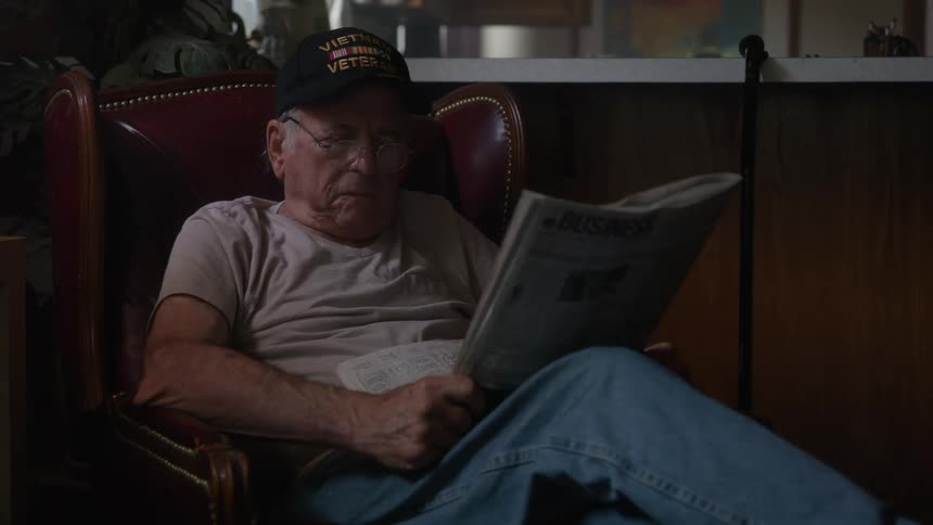 An elderly man wearing a hat is sitting in a lounge chair reading a newspaper when someone in front of him gets up making him complain about something