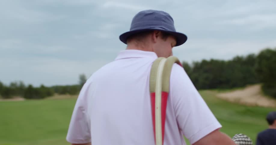 A man walks down a golf course following other people carrying a pink golf bag while smiling and talking to someone behind him