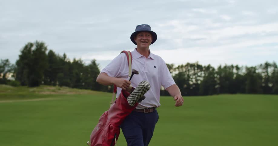 Man with hat pink polo shirt dark blue pants and red staff bag walks in the golf course smiling