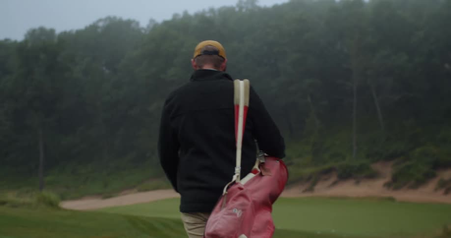 Golfer with cap coat pants and red golf bag walks across golf course