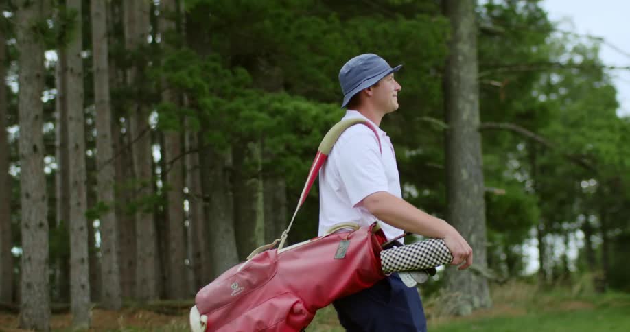 Man carries golf bag while walking over course past conifer grove