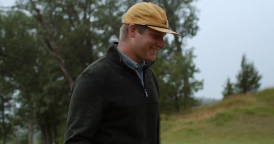 Man with yellow cap black sweater and brown pants walks uphill in golf course laughing