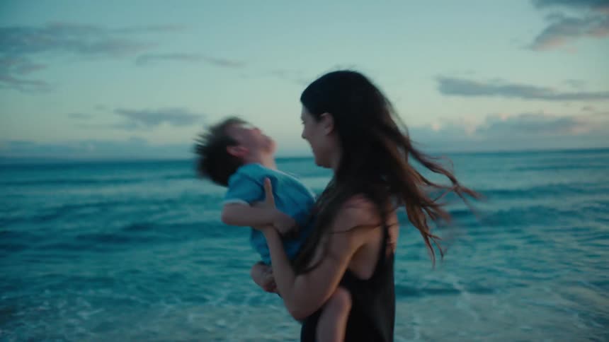 A woman spins around playfully while holding a young boy in her arms on a beach during the day