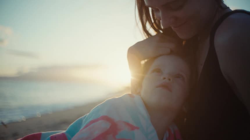 A toddler is held by his mother on a beach as she caresses his head and he looks up both of them smiling
