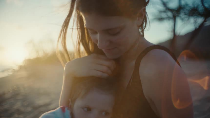 A woman on a beach holds her toddler in her arms and caresses his hair as she stairs at the ocean