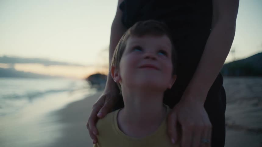 Mother steps up behind son to put hands on his shoulders and he smiles up at her as waves roll onto beach